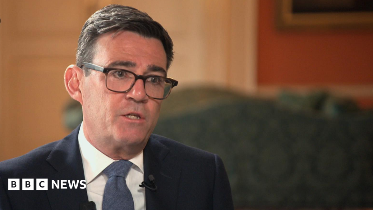 Andy Burnham wears a dark suit sitting on a chair speaking with Roger Johnson who wears a dark suit. They are both sitting on red chairs.