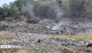 An image of a large pile of industrial waste in a field. The waste has small plumes of smoke coming from it, having recently been extinguished by fire crews.