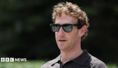 A close-up of Mark Zuckerberg, wearing a set of Meta Ray-Bans and brown top, walking through a park.