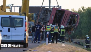 A crane winches an overturned bus up on the side of the road as firefighters look on.