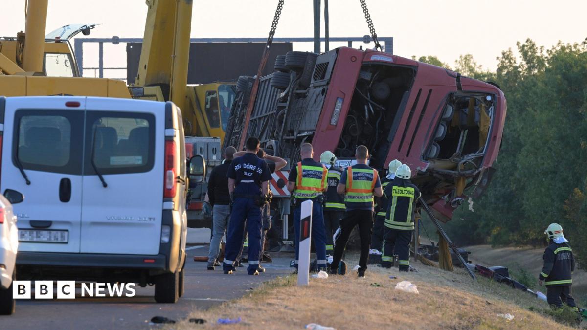 A crane winches an overturned bus up on the side of the road as firefighters look on.