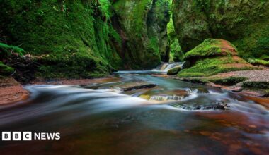 A long exposure shot of water flowing down a steep gorge. A large rock can be seen in the foreground and steep green covered rocks can be seen in the background