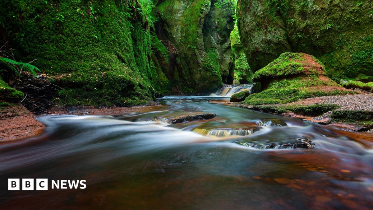 A long exposure shot of water flowing down a steep gorge. A large rock can be seen in the foreground and steep green covered rocks can be seen in the background
