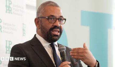 James Cleverly wears a suit and holds a microphone