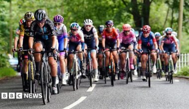 A group of cyclists in colourful racing kits ride closely together on a tree-lined road during a road race, with several riders competing for the lead.