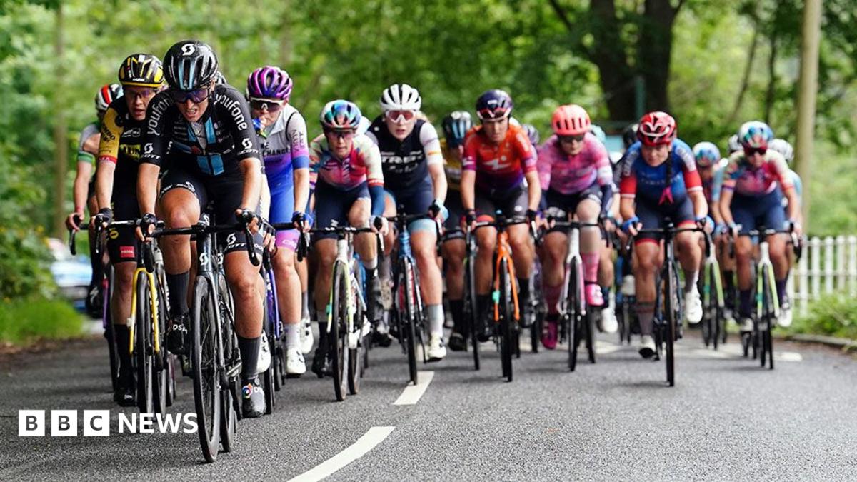 A group of cyclists in colourful racing kits ride closely together on a tree-lined road during a road race, with several riders competing for the lead.