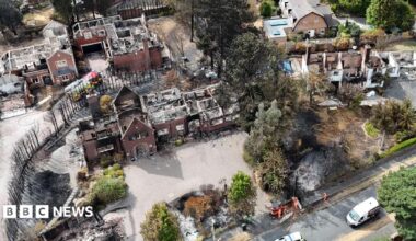 An aerial image shows several houses destroyed by fire, with their roofs gone and wreckage visible within. There is a line of dead trees next to the houses and areas of scorched earth. A fire engine is parked by one of the houses.