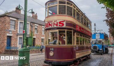 Visitors travel on a vintage electric tram in the 1900s village at Beamish Museum. There is a row of terraced houses on a cobbled street. Two double-decker public transport vehicles from different eras are in shot. The front one is a maroon and beige tram with a curved front and a large advert for Binns between the two floors. The rear one has more of the appearance of an old trolleybus or lorry, with a cab for the driver, an exposed radiator and distinct tyres. It is bright blue and open-topped.