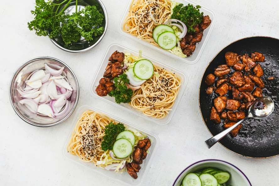 Image of a few food containers filled with stir-fried noodle salads on a marble counter, alongside a pan with chicken and bowls of vegetables for meal planning.