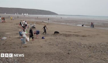 A wide sandy beach at low tide stretches towards the sea, with shallow pools of water scattered across the exposed sand. In the foreground, a large sandcastle creation featuring circular and curved walls has been built on the beach, surrounded by brightly coloured buckets, spades and other beach toys. Several people are walking and playing nearby, including children running across the sand and a person standing close to the sand structure.
