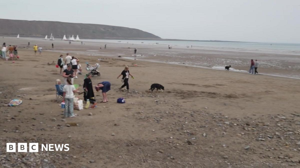 A wide sandy beach at low tide stretches towards the sea, with shallow pools of water scattered across the exposed sand. In the foreground, a large sandcastle creation featuring circular and curved walls has been built on the beach, surrounded by brightly coloured buckets, spades and other beach toys. Several people are walking and playing nearby, including children running across the sand and a person standing close to the sand structure.