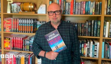 A man standing in front of giant book case full of books, he is wearing a checked shirt and black glasses. He is holding a book in his hands