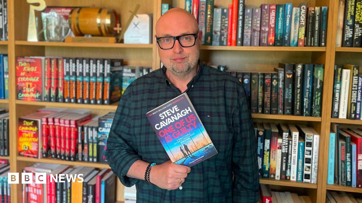 A man standing in front of giant book case full of books, he is wearing a checked shirt and black glasses. He is holding a book in his hands