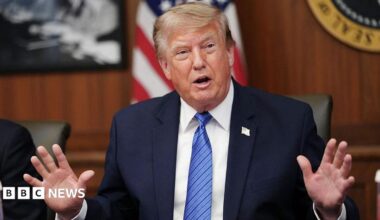 Donald Trump holds up his right fist while wearing a white shirt and red tie.