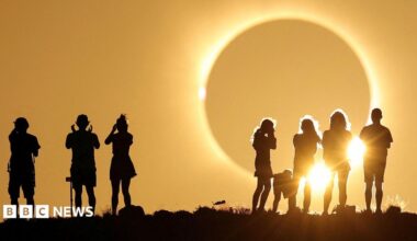 Crowds of mixed genders and ages stand on a grassy hilltop with the columns of the National Monument visible in the background. A group of friends at the front can be seen wearing eclipse glasses.
