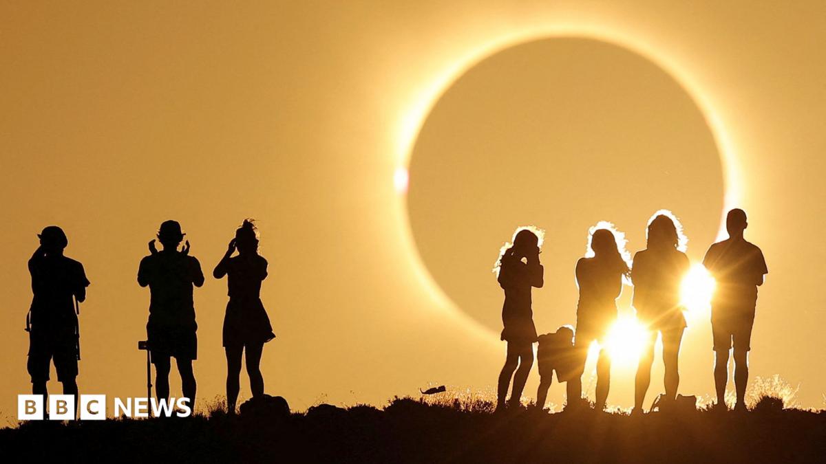Crowds of mixed genders and ages stand on a grassy hilltop with the columns of the National Monument visible in the background. A group of friends at the front can be seen wearing eclipse glasses.