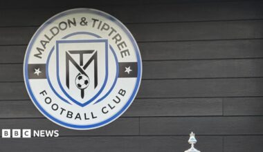 A blue sign reads "Home of Maldon & Tiptree FC". Police cars and vans are parked beyond it.