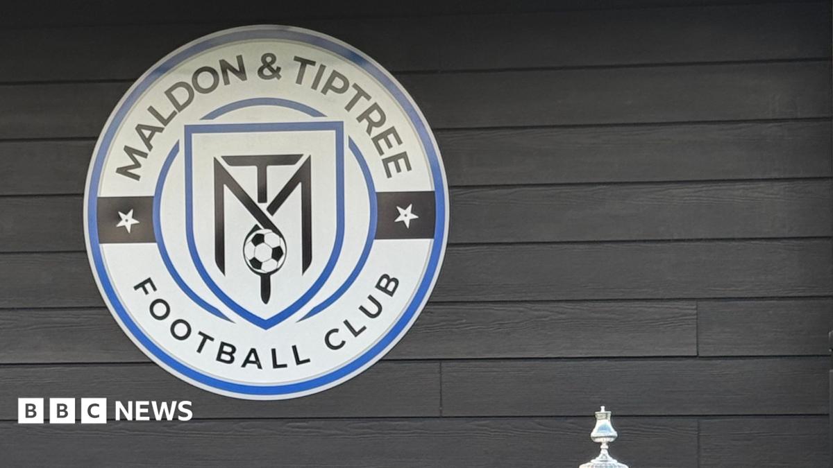 A blue sign reads "Home of Maldon & Tiptree FC". Police cars and vans are parked beyond it.