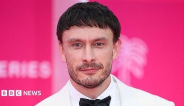 Richard Gadd has short, dark hair and a short beard and is wearing a white suit jacket, white shirt and black bow tie. He is standing in front of a pink background