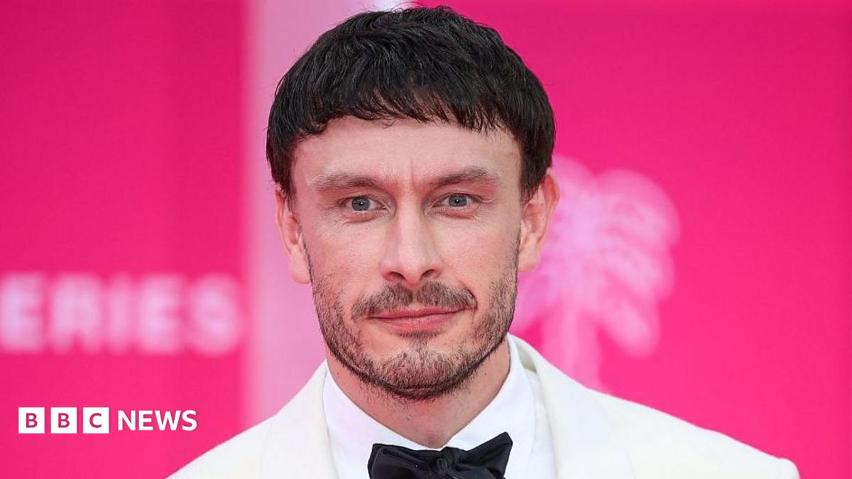 Richard Gadd has short, dark hair and a short beard and is wearing a white suit jacket, white shirt and black bow tie. He is standing in front of a pink background