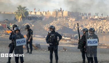 Spanish armed police with riot shields on Trampolin beach, Ceuta, as anti-migrant protesters set fire to a migrant camp there. Their faces are blurred. Smoke can be seen rising in the air.