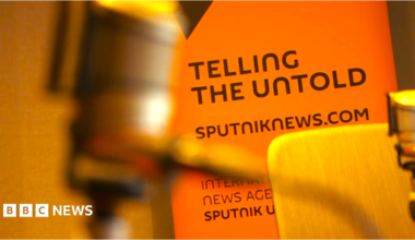 A SputnikNews.com bill board with an out-of-focus microphone in the extreme foreground. We can see the wooden chair of a radio studio. The billboard is a deep orange with black lettering saying "Telling the Untold", and sputniknews.com.