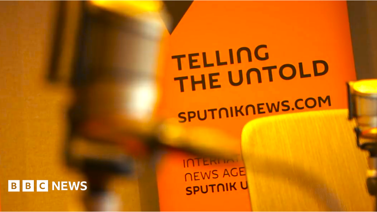 A SputnikNews.com bill board with an out-of-focus microphone in the extreme foreground. We can see the wooden chair of a radio studio. The billboard is a deep orange with black lettering saying "Telling the Untold", and sputniknews.com.