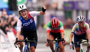 Grace Brown of team FDJ Nouvelle-Aquitaine Futuroscope celebrates as she crosses the line to win stage four of The Women's Tour from Wrexham to Welshpool on 2022. She is wearing a blue and white cycling suit, glasses and a white helmet. Her arms is raised in a fist as she rides the bike.