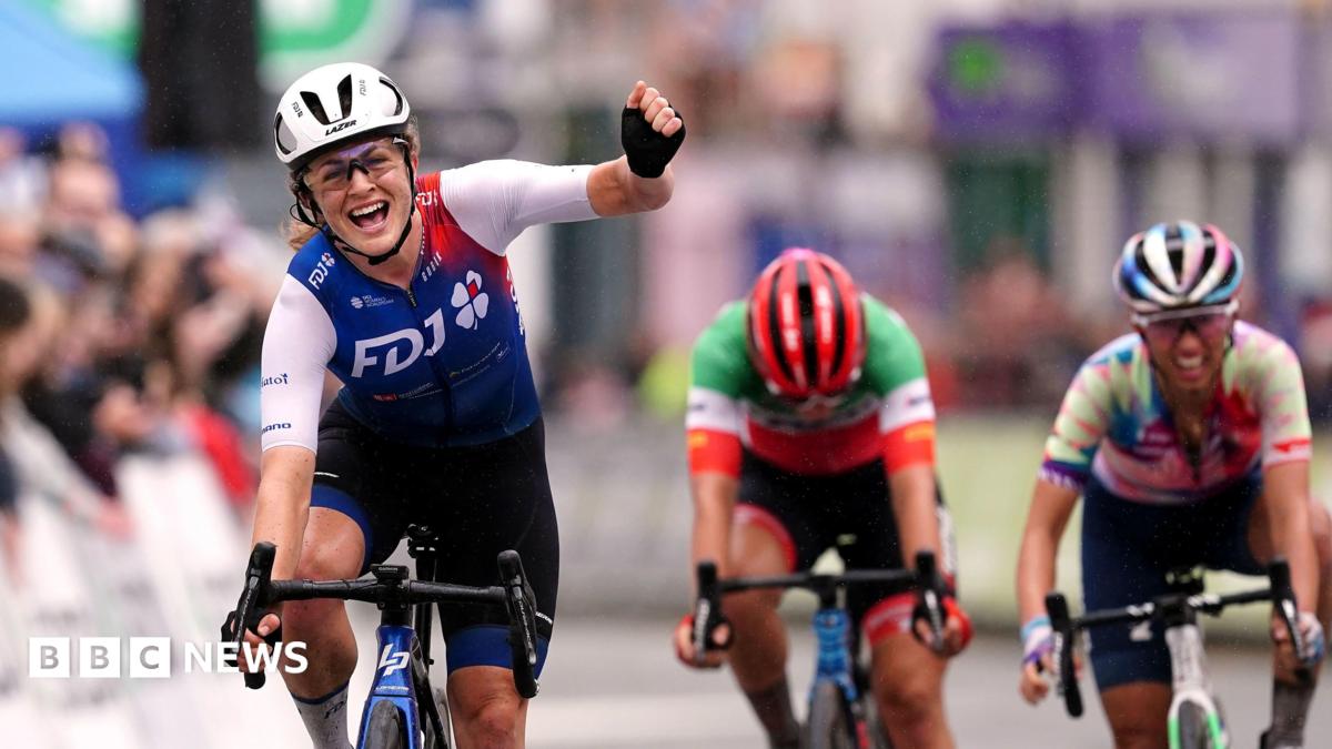 Grace Brown of team FDJ Nouvelle-Aquitaine Futuroscope celebrates as she crosses the line to win stage four of The Women's Tour from Wrexham to Welshpool on 2022. She is wearing a blue and white cycling suit, glasses and a white helmet. Her arms is raised in a fist as she rides the bike.
