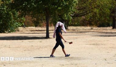 Person walking across a very dry and brown park with a towel over their head, providing shelter from the hot weather