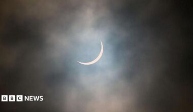 The sun is pictured during a partial solar eclipse. The sky is dark and cloudy. The sun is a thin arc in the sky.