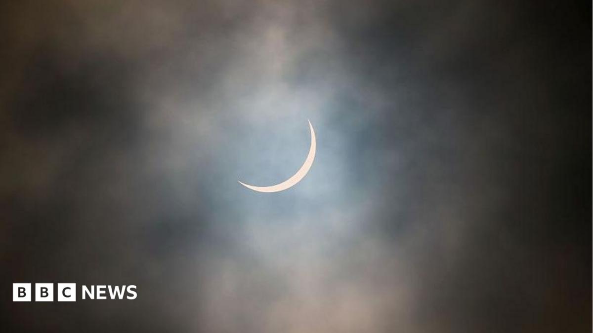 The sun is pictured during a partial solar eclipse. The sky is dark and cloudy. The sun is a thin arc in the sky.