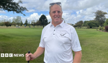 Andy Wilkins - a man with grey hair and sunglasses on his head - smiling. He is wearing a white polo shirt with an emblem of a golf club. He is holding a golf club. He's standing on the golf green with golfers behind him and a flag and some trees