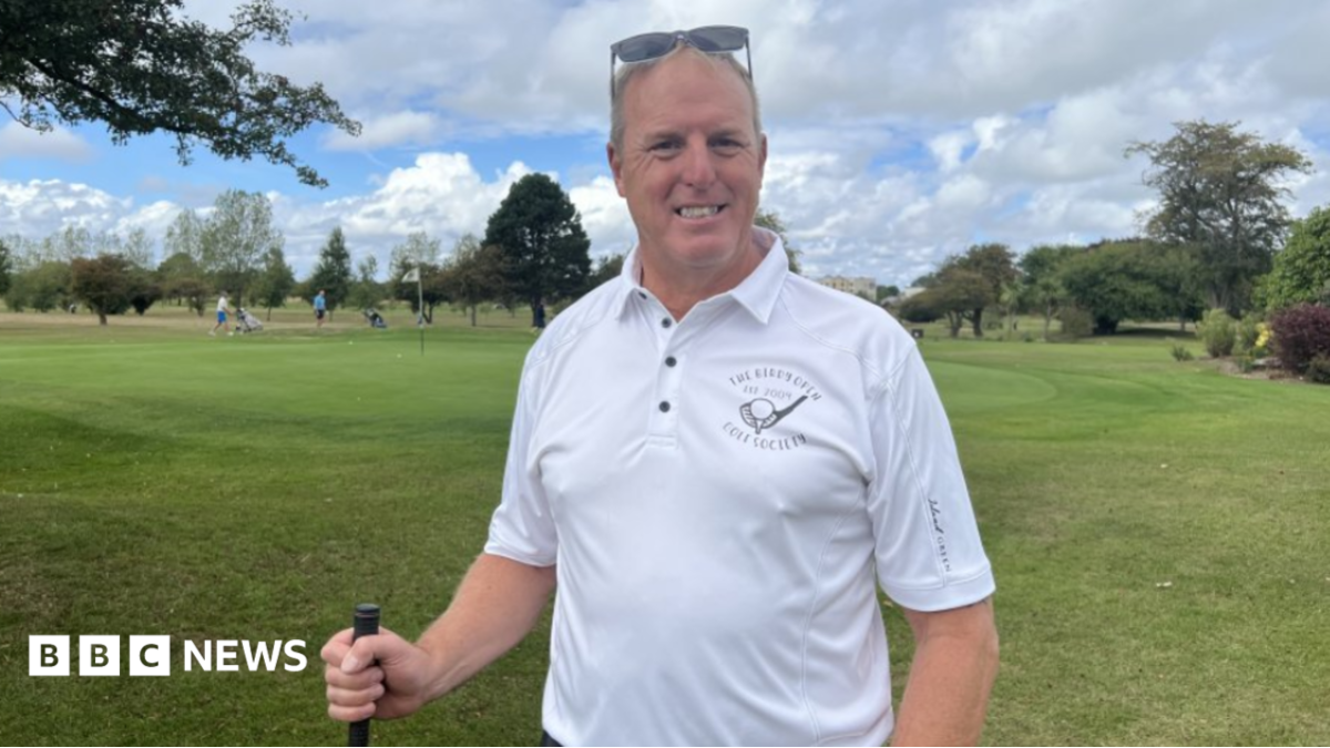 Andy Wilkins - a man with grey hair and sunglasses on his head - smiling. He is wearing a white polo shirt with an emblem of a golf club. He is holding a golf club. He's standing on the golf green with golfers behind him and a flag and some trees