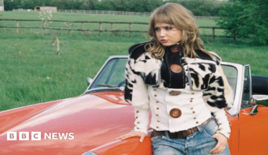 Alexandra Burch during a photoshoot of her cowhide jacket. She is leaning on a red car parked by a green field.