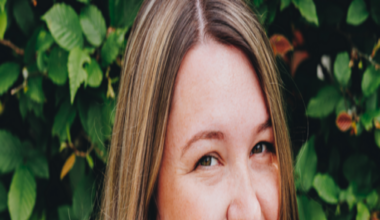 A headshot of a woman with blonde hair stood in front of bushes outside.