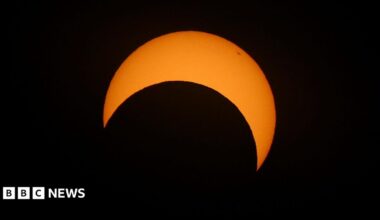 The sun is pictured during a partial solar eclipse. The sky is dark and cloudy. The sun is a thin arc in the sky.