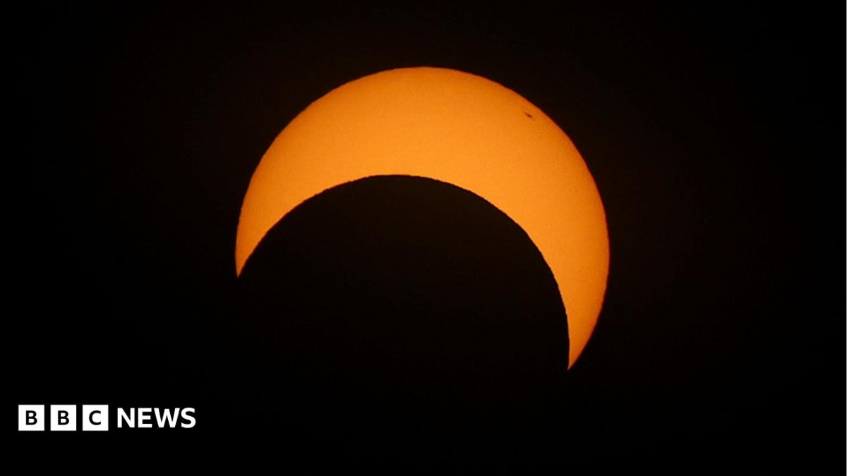 The sun is pictured during a partial solar eclipse. The sky is dark and cloudy. The sun is a thin arc in the sky.