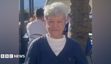 A woman with short grey hair sits at an outdoor café, wearing a navy cardigan over a light-coloured top. A tree trunk is beside the table, with other customers seated in the background.
