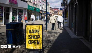 A high street scene in which there is a yellow sign with "Vape Shop Open" in black text. The sign is next to a bin. Pedestrians are in the background and there are shops either side of the walkway.