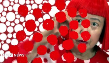 Yayoi Kusama looks at the camera wearing round glasses and a red top with white polka against a backdrop of polka dot tentacles for her exhibition in Hong Kong in 2012.