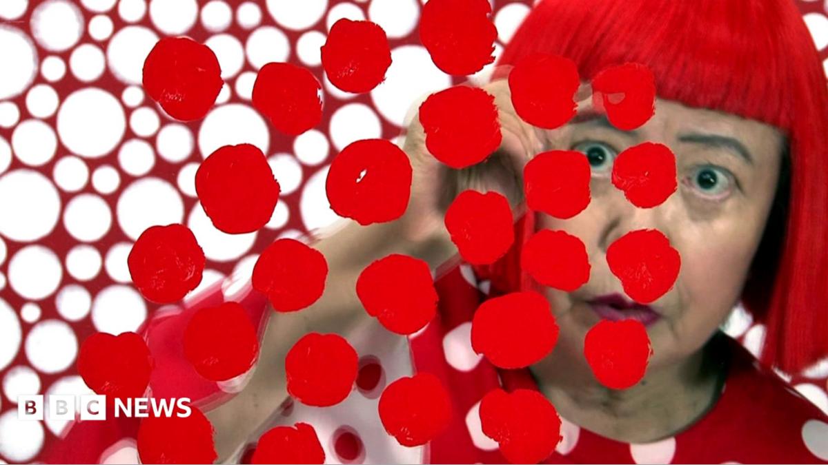 Yayoi Kusama looks at the camera wearing round glasses and a red top with white polka against a backdrop of polka dot tentacles for her exhibition in Hong Kong in 2012.