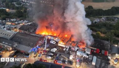 An aerial view of a huge seat of fire across a large industrial complex with a plume of smoke rising high