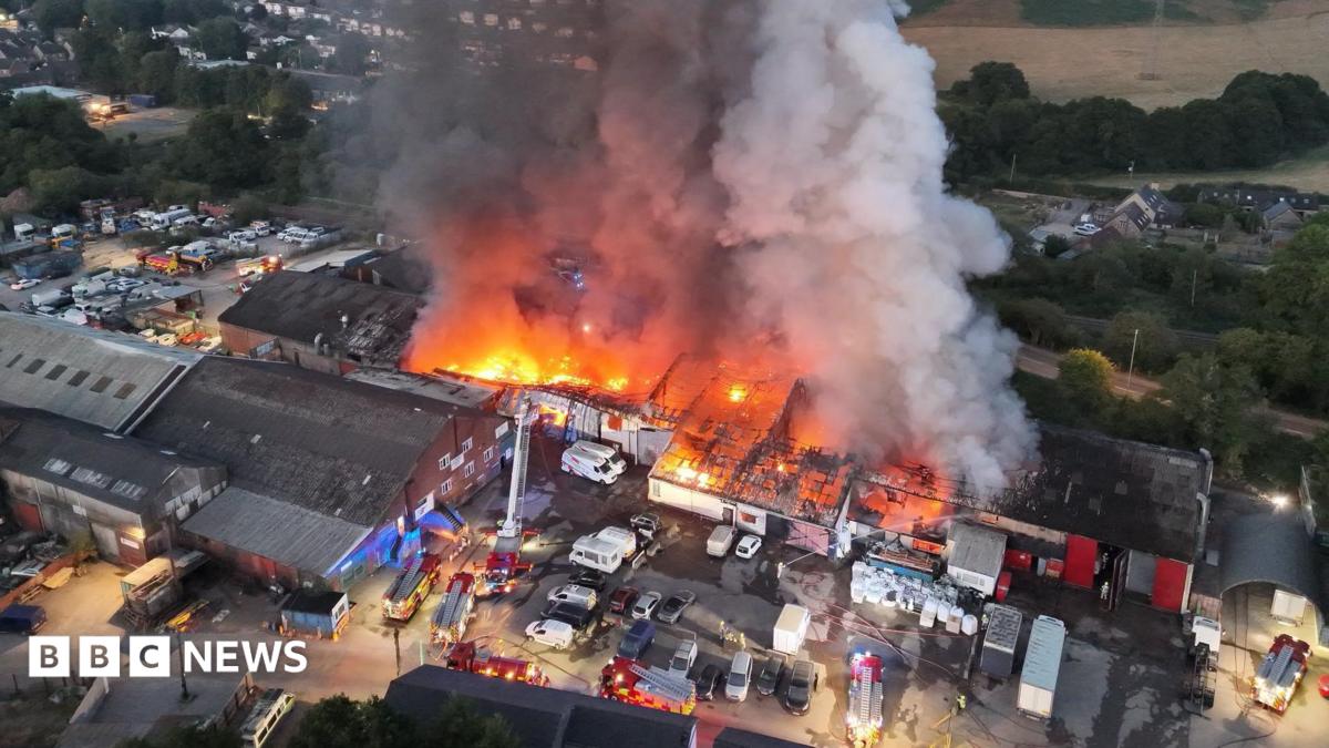 An aerial view of a huge seat of fire across a large industrial complex with a plume of smoke rising high
