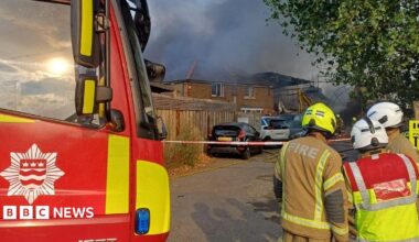 Firefighters stand beside a London Fire Brigade engine as thick black smoke rises from a large fire behind nearby buildings in Mitcham. Emergency crews have cordoned off the area while tackling the blaze at a workshop and scaffolding yard.