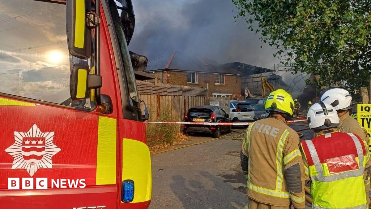 Firefighters stand beside a London Fire Brigade engine as thick black smoke rises from a large fire behind nearby buildings in Mitcham. Emergency crews have cordoned off the area while tackling the blaze at a workshop and scaffolding yard.