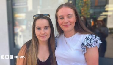 Beatrice and Amelia stand side by side outdoors, looking towards the camera. Amelia is wearing a dark sleeveless top with sunglasses resting on their head, while Beatrice is wearing a white top with patterned ruffled sleeves. The background is softly blurred and shows a modern building entrance.