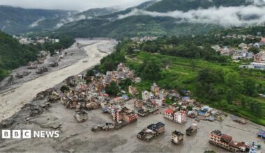 Aerial view of a flood-hit town in a valley in Nepal, with muddy water and debris surrounding damaged buildings beside a swollen river.