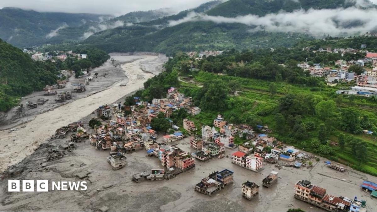Aerial view of a flood-hit town in a valley in Nepal, with muddy water and debris surrounding damaged buildings beside a swollen river.