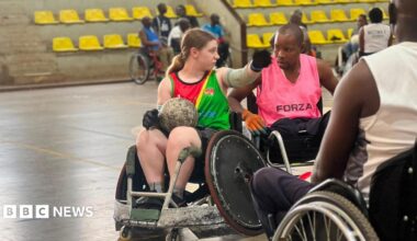 Sarah Burrows (left) in conversation with a man in a sports hall. They are both in wheelchairs. Sarah has a round ball in her lap. She has brown hair, tied back, and is wearing a red and green top. The man next to her has short black hair and is wearing a pink top.